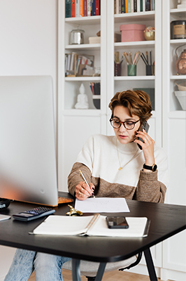 woman on phone wearing glasses in home office in front of book shelf