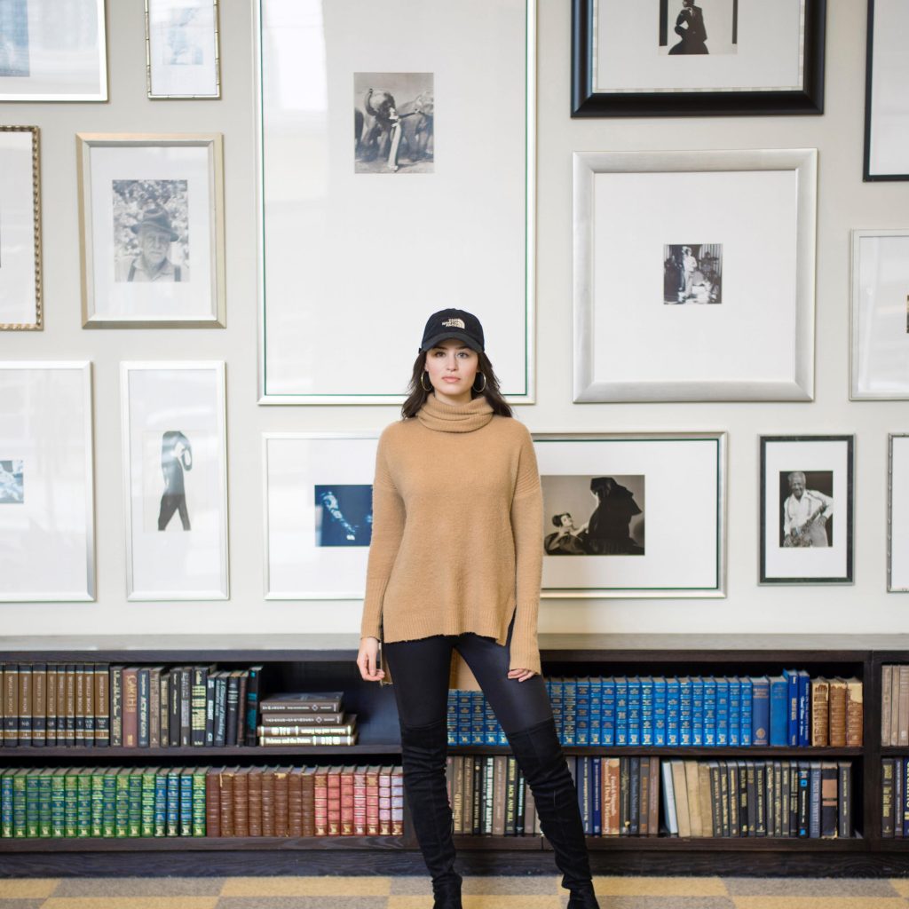 Creativity Takes Courage - woman in a hat and yellow sweater standing in front of a wall of art and books