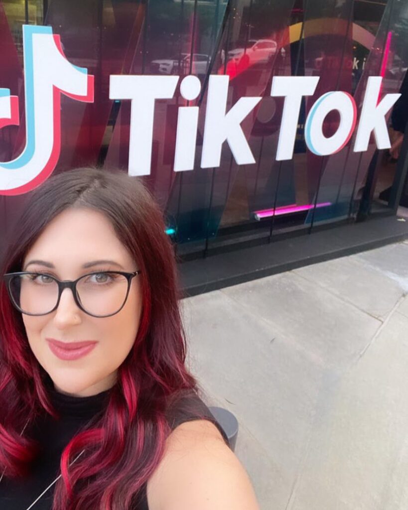 Jennifer Paukman stands in front of TikTok sign. She has red hair and wears glasses.